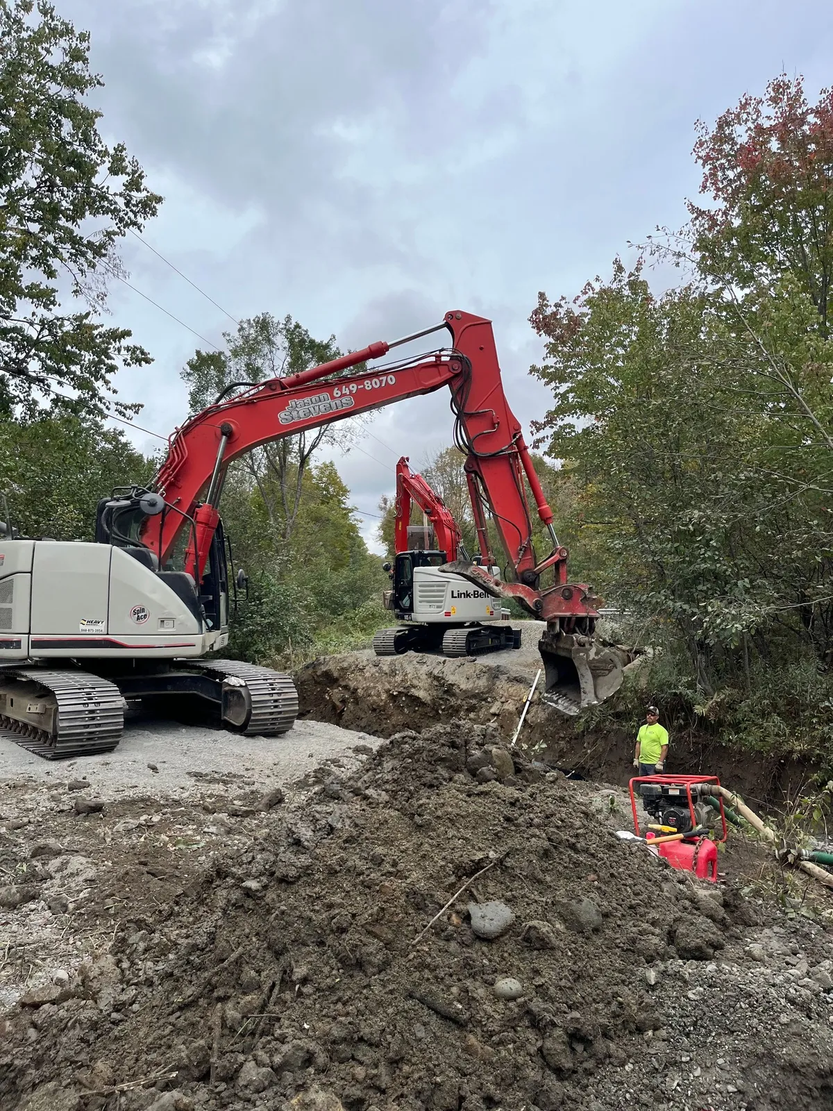 Two red excavators working a dirt site with piles of soil and a worker in a high-visibility vest.