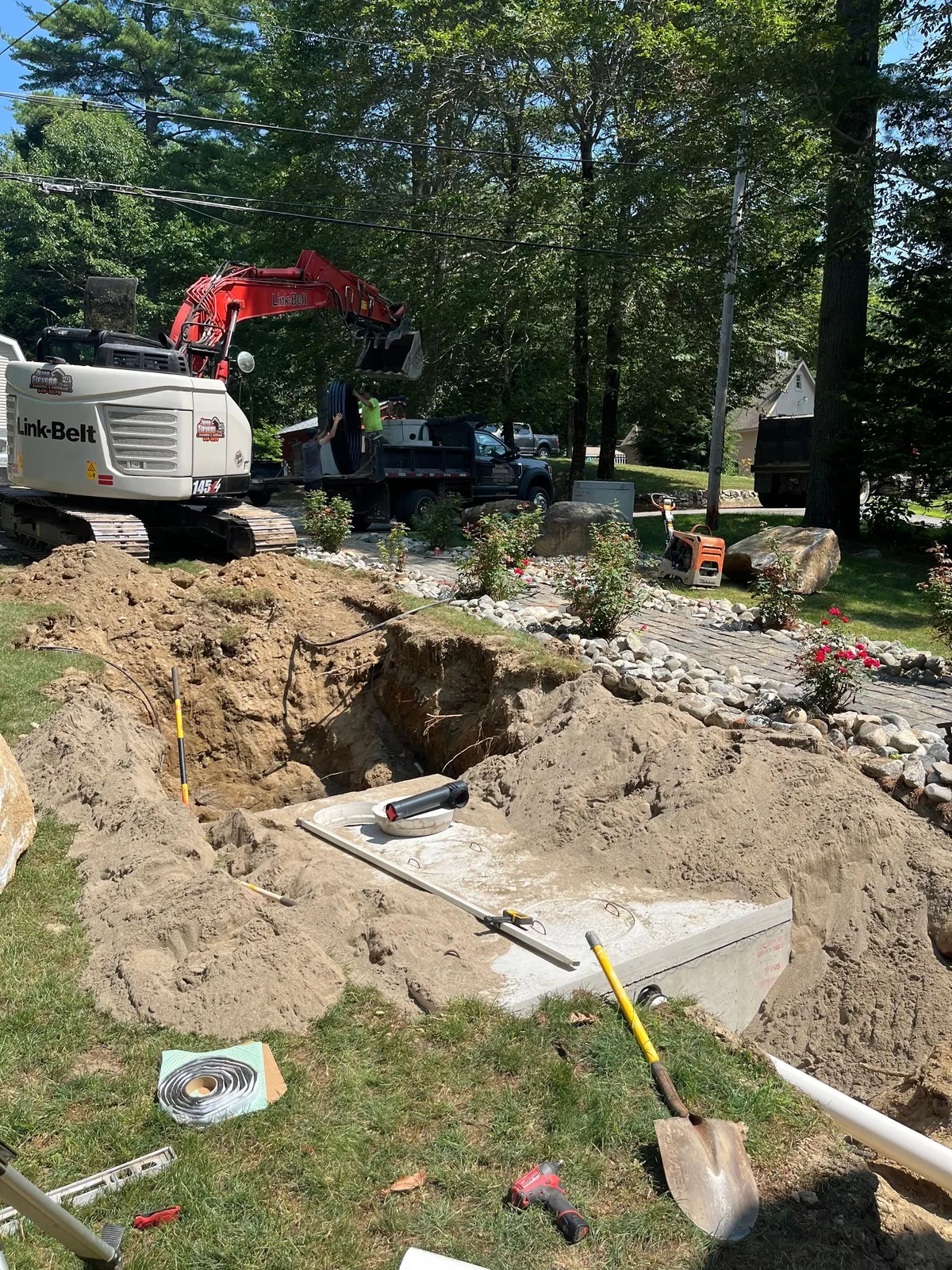 A red excavator beside an open trench holding a concrete septic tank on a grassy Maine property.