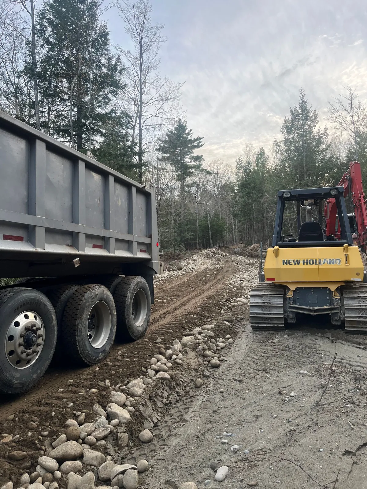 A crawler loader and dump truck on a gravel access path in the woods.